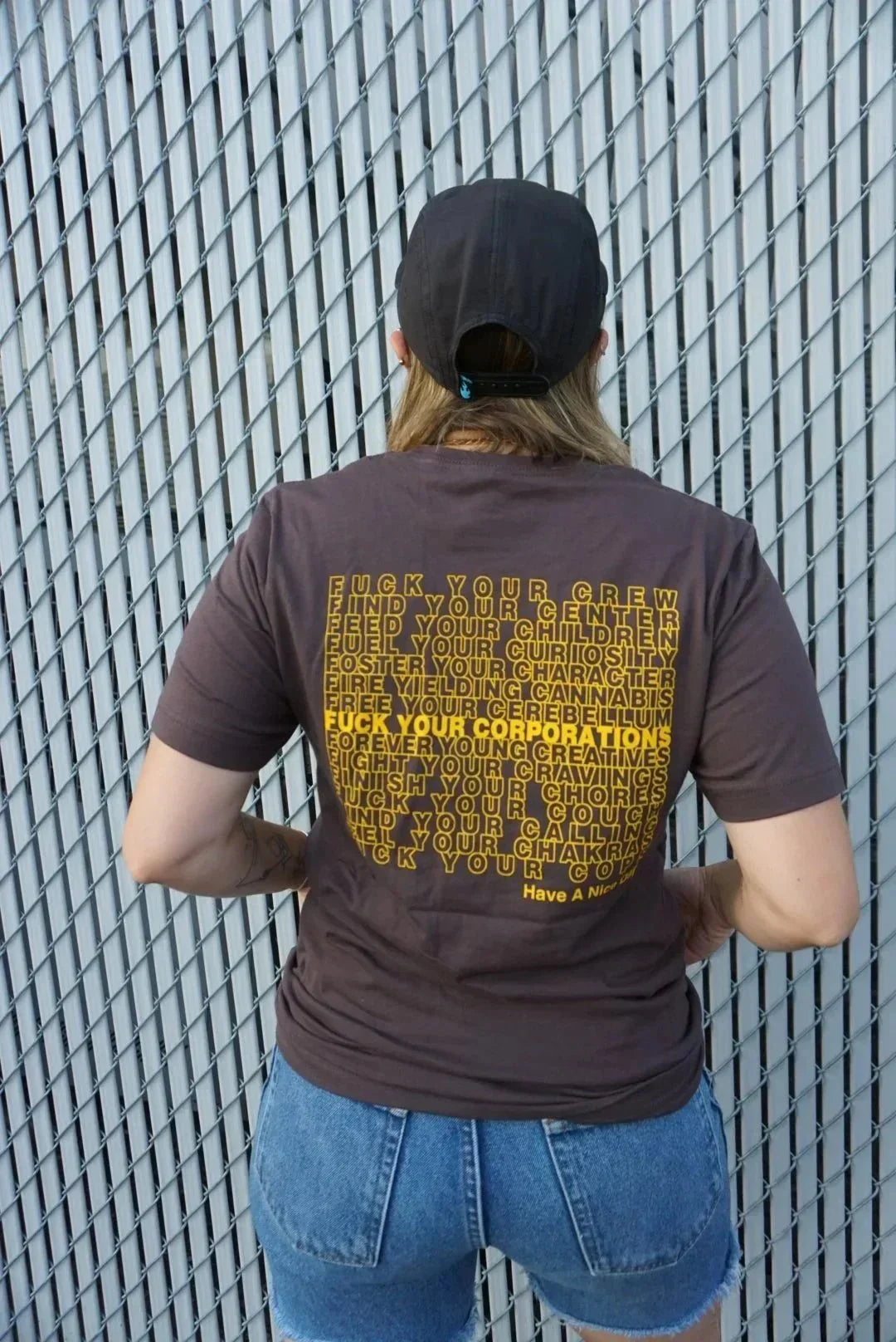 Person wearing black cap, brown graphic tee with bold yellow text, and denim shorts, standing by a metal fence.
