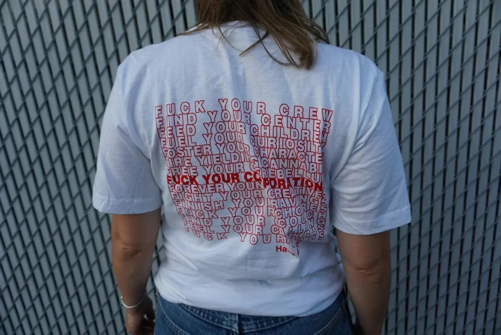 Person wearing a white graphic t-shirt with bold red text on the back, standing by a chain-link fence.
