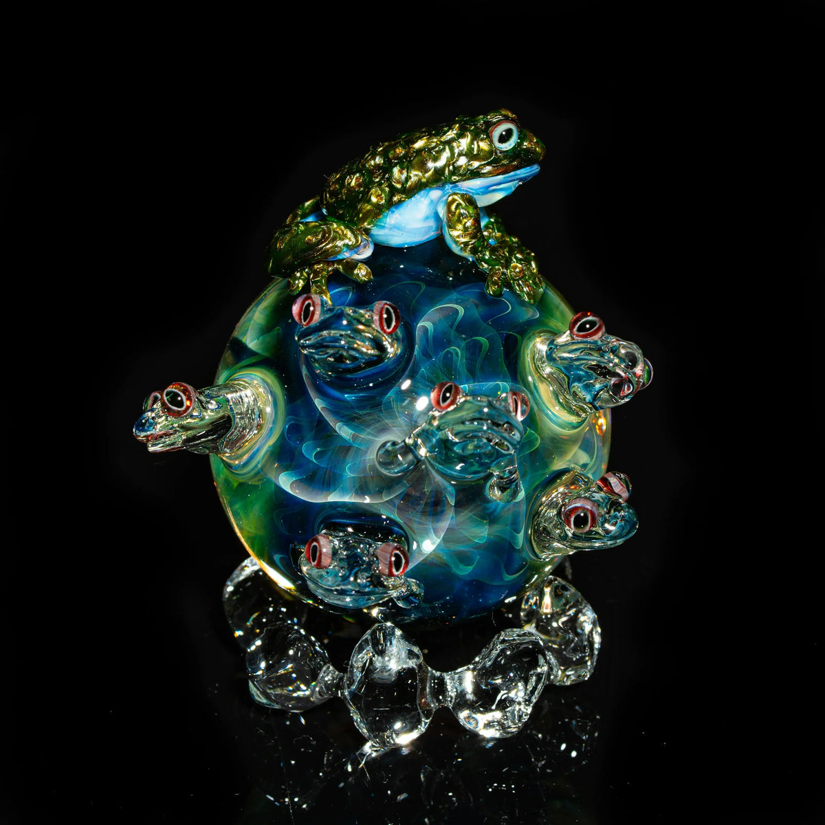 Artistic glass sculpture featuring frogs on a colorful orb, displayed on a reflective surface