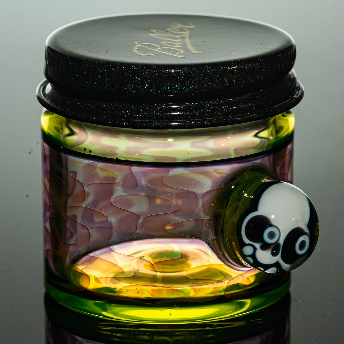 Colorful heady glass jar with black lid and decorative skull accent, reflective surface