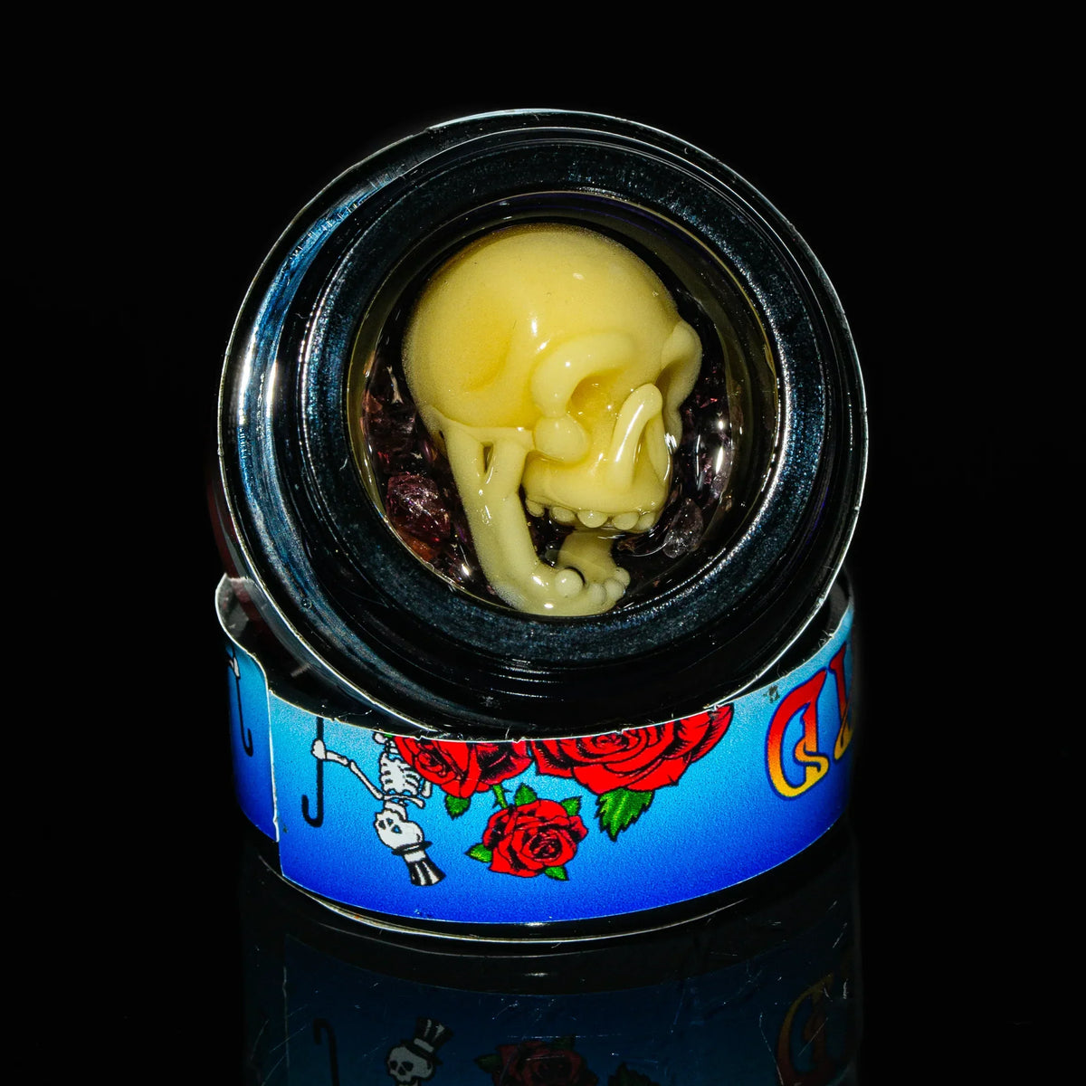 Yellow glass skull art inside a jar with blue and red rose skeleton design on base
