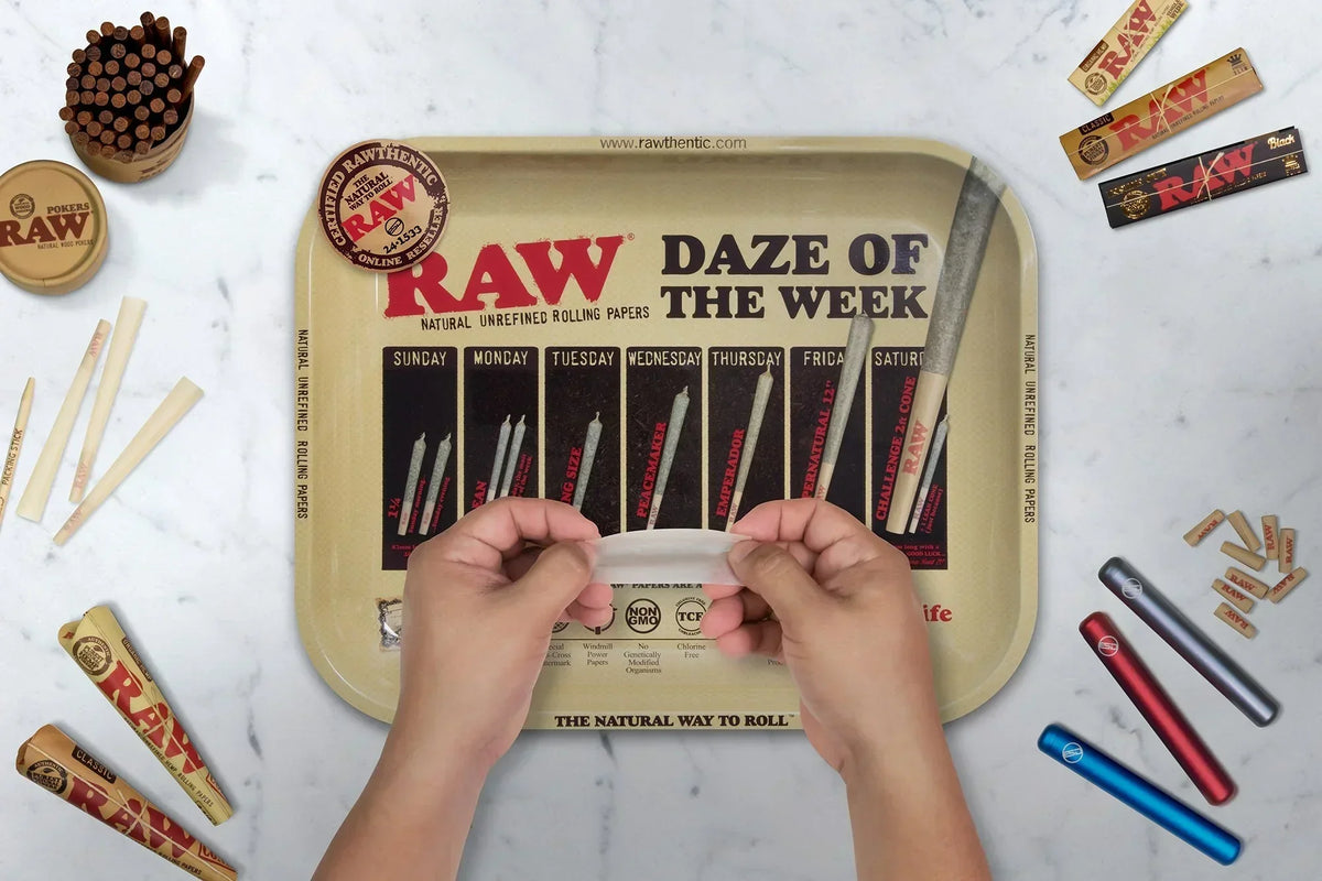 Large RAW rolling tray with Daze of the Week design, hands holding rolling paper, smoking accessories