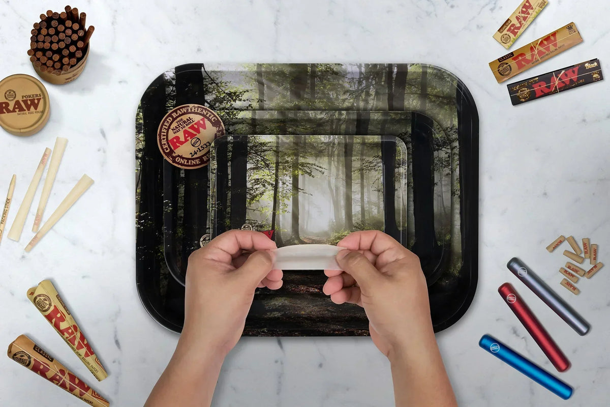 Hands holding rolling paper above forest-themed rolling tray with papers, cones, tubes, and filters on marble surface.