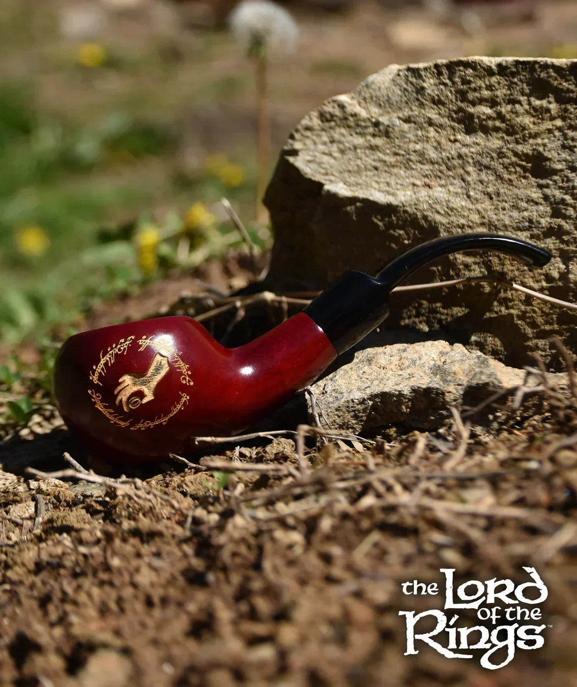 Lord of the Rings themed wooden pipe with gold inscription, outdoors on soil by a rock