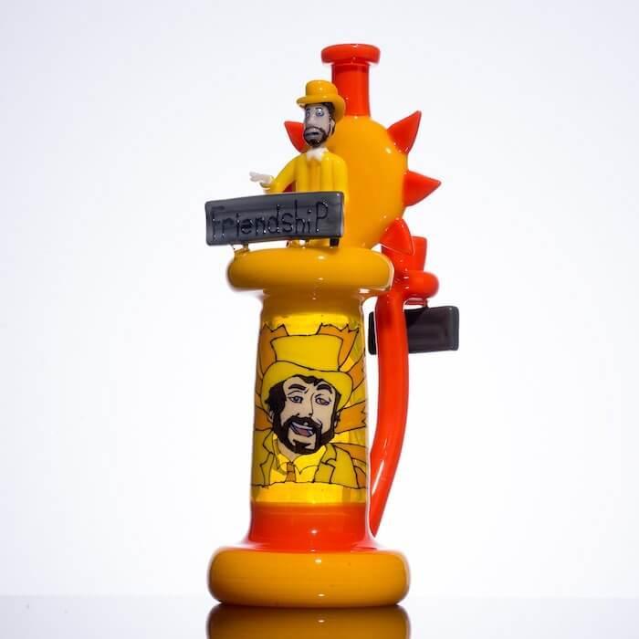 Vibrant yellow and orange glass pipe with cartoon figure, 'friendship' sign, and sunburst design