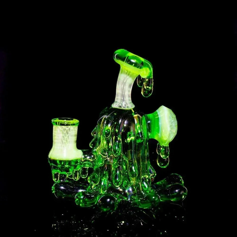 Hand-blown green glass mini rig with splatter design and matching cap on black background.