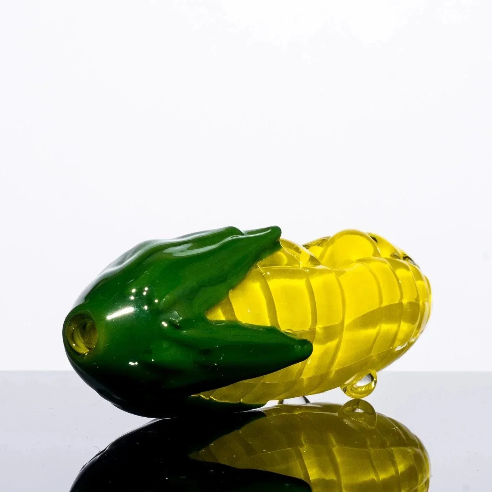 Hand-blown glass corn cob pipe with green husk, yellow kernels, on reflective surface