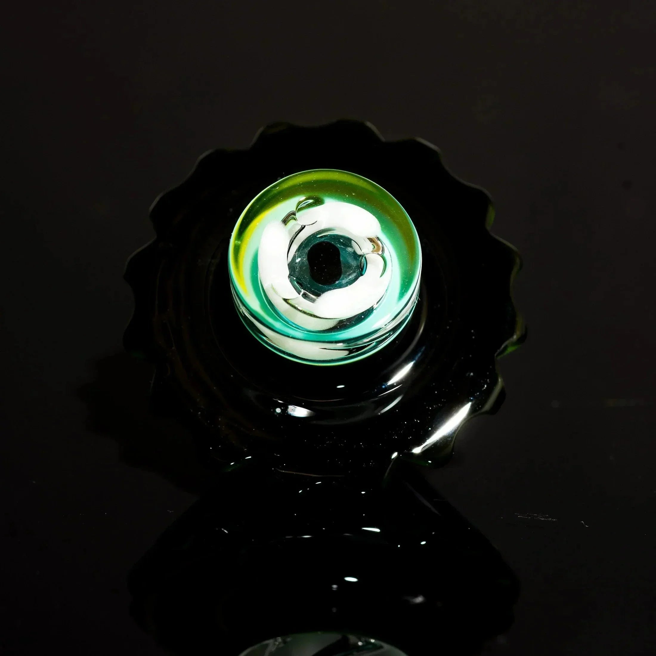 Handblown glass pipe bowl with green and white swirls, black scalloped base, on dark reflective surface