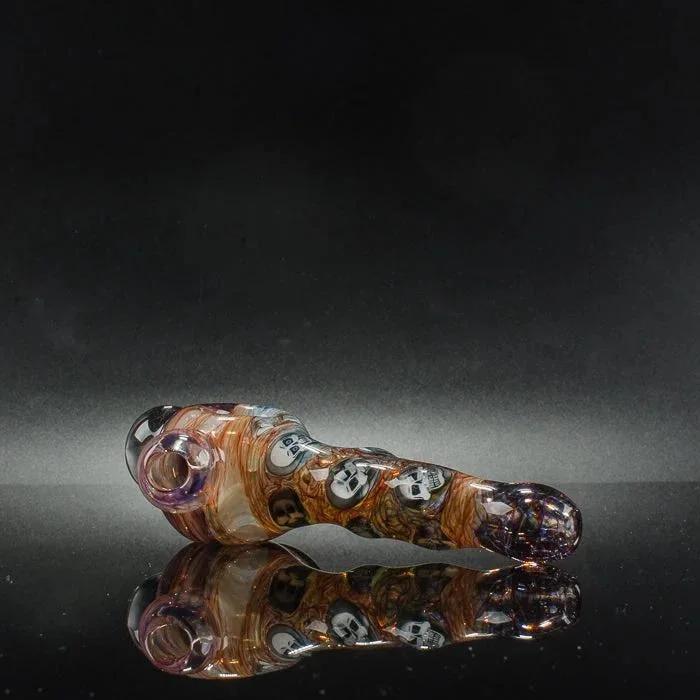 Handblown glass pipe with skull motifs, amber swirls, and purple accents on reflective black surface