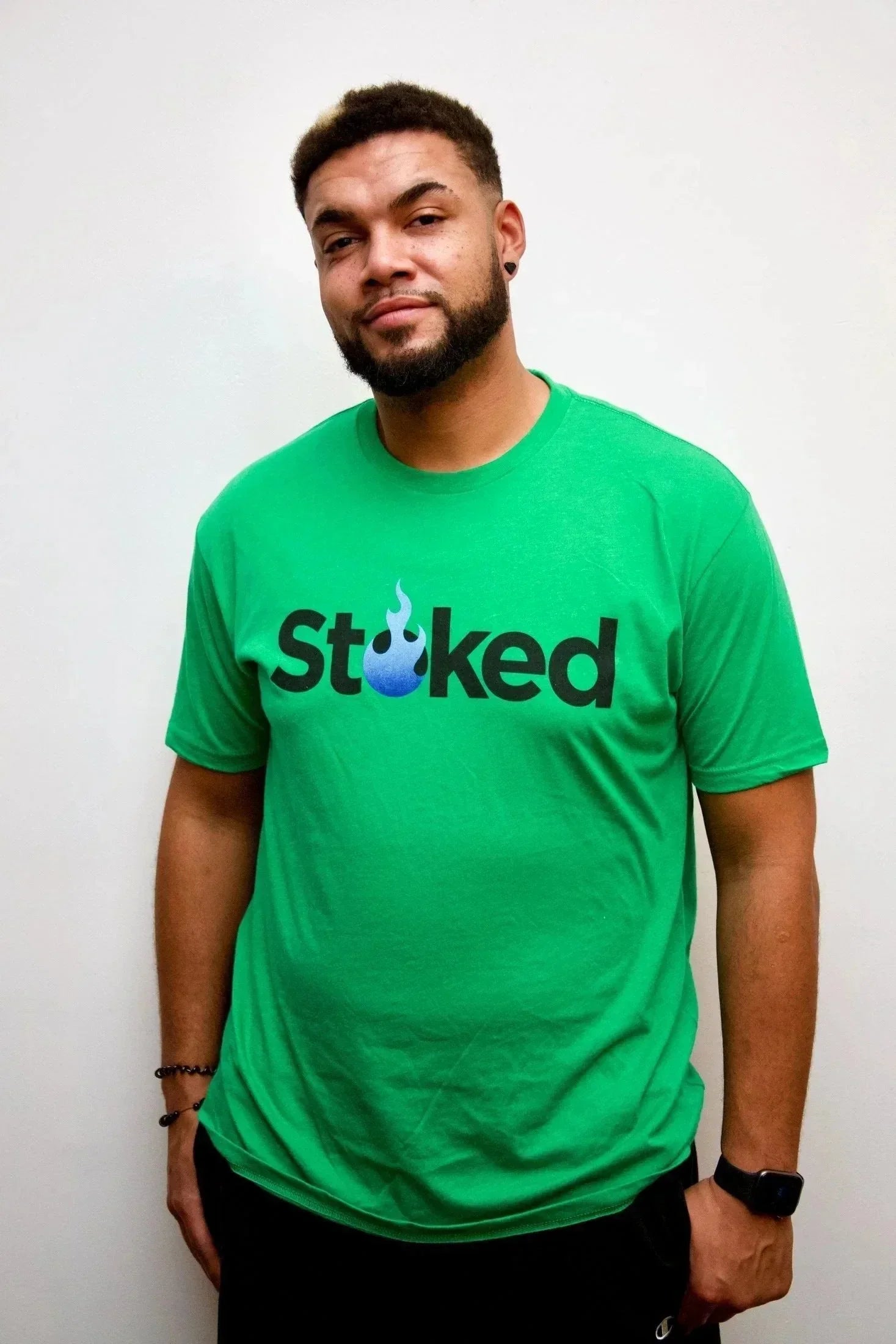 Man wearing green Stoked t-shirt with blue flame logo on plain background