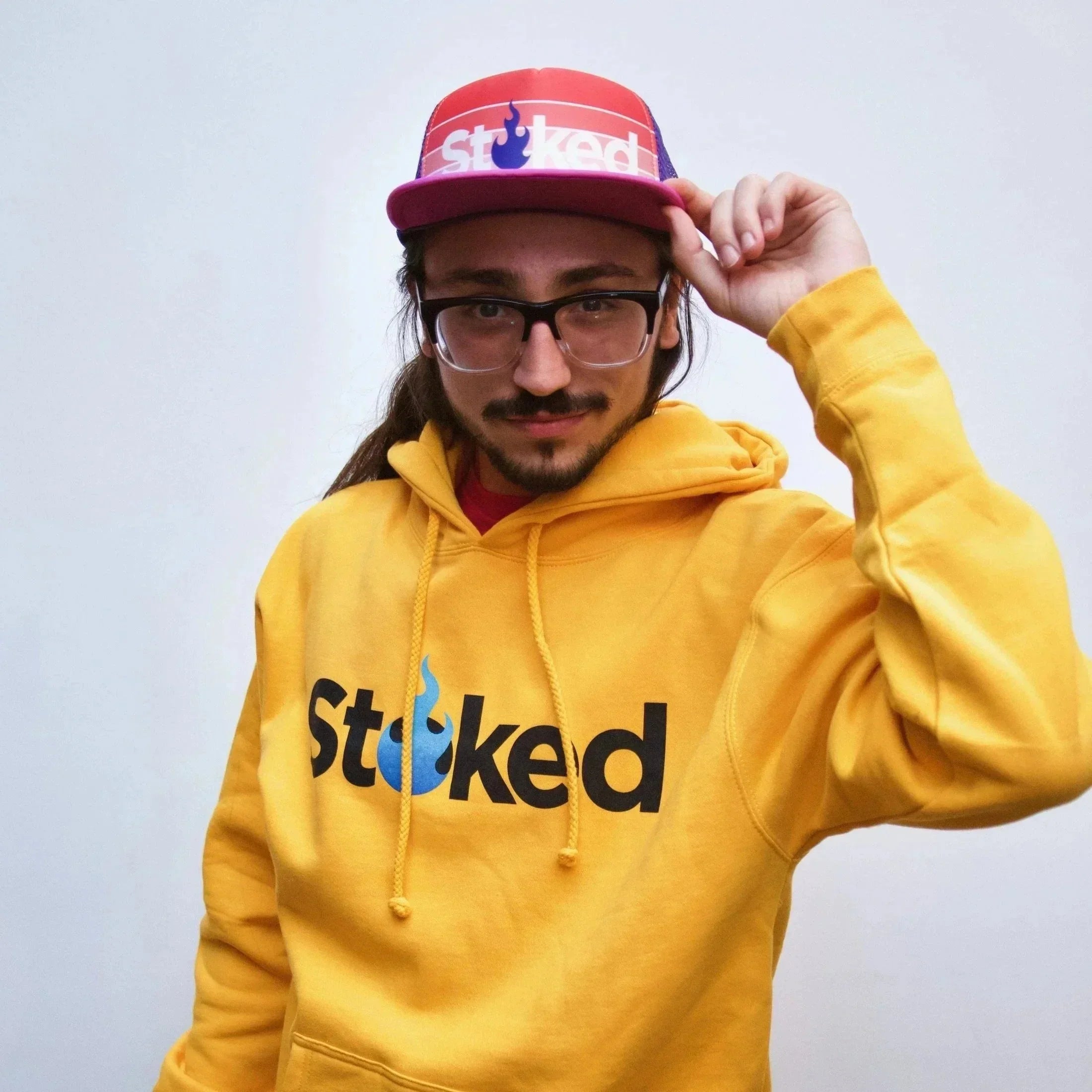 Man in yellow hoodie with 'Stoked' logo and red cap, standing against white background
