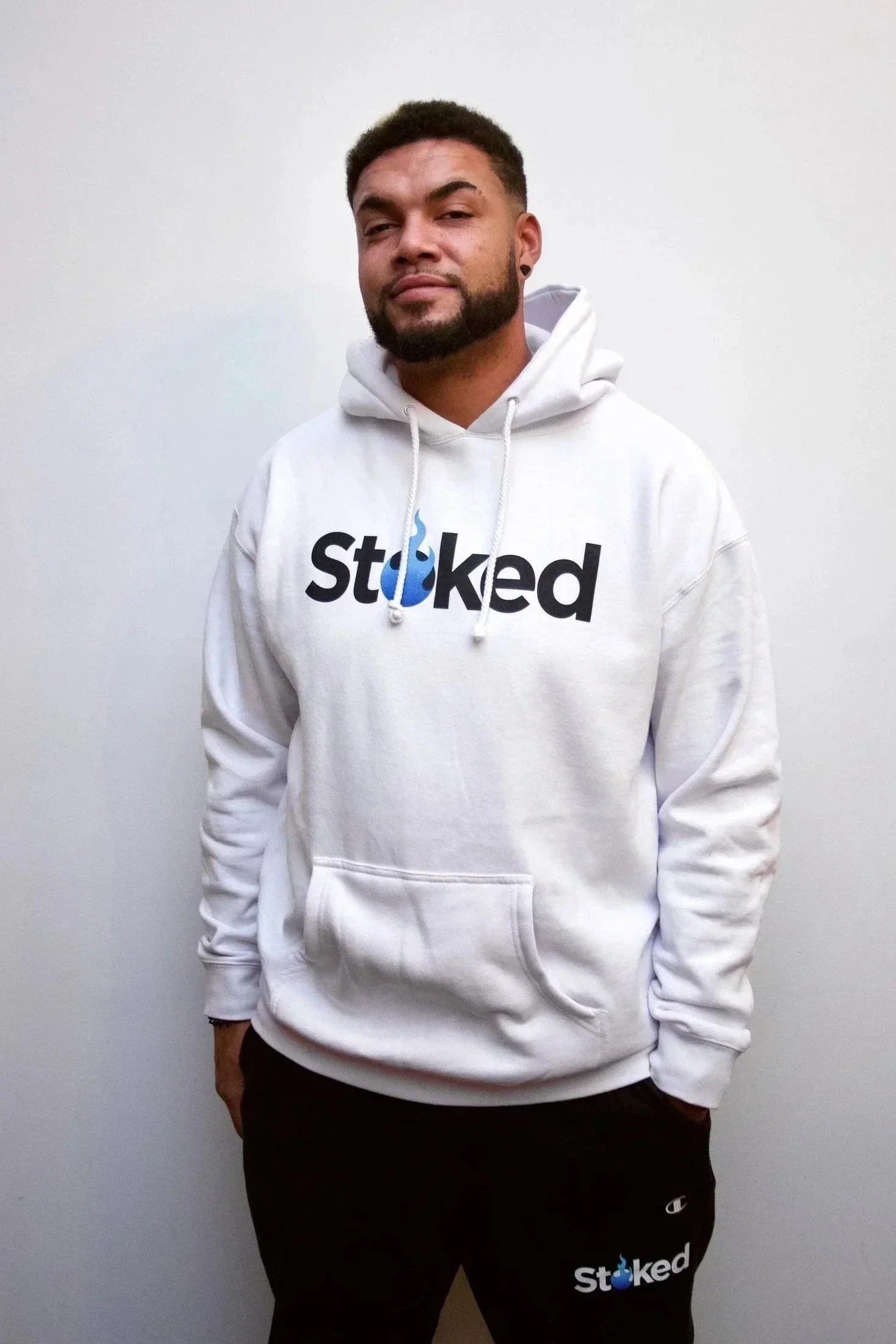 Man wearing white Stoked logo hoodie and black joggers against plain background