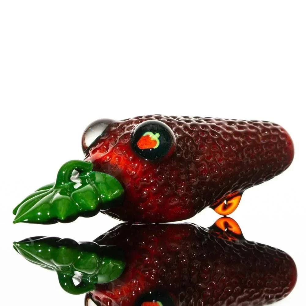 Handcrafted glass pipe shaped like a strawberry with green leaf detail, reflected on surface.