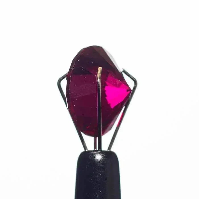 Deep red faceted gemstone held in black tweezers against white background