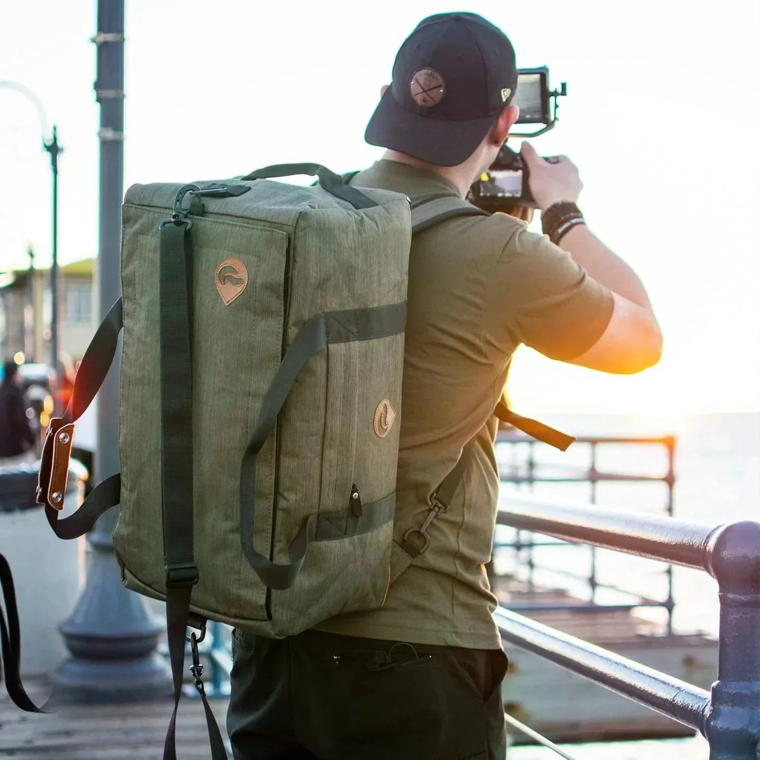 Man wearing a large green hybrid backpack outdoors at sunset, taking a photo by the water