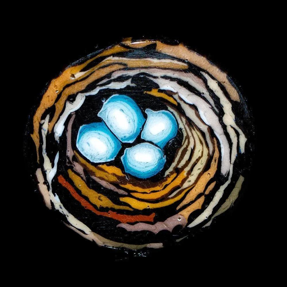 Glass coin art featuring a bird’s nest with three blue eggs on a black background.