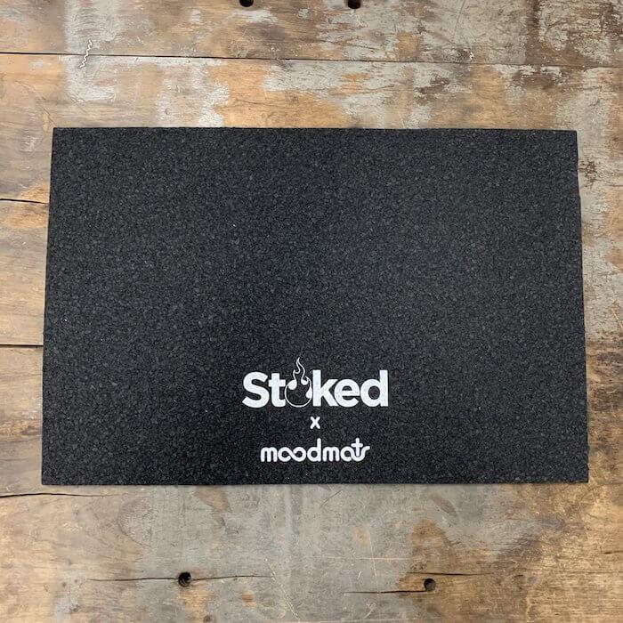 Stoked x moodmat black grip mat on rustic wood, ideal for glass pipe or vaporizer use