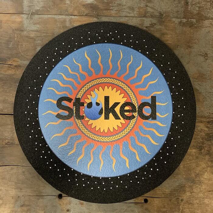 Stoked 17 inch circular mood mat on wood background with Stoked sun logo in orange and blue