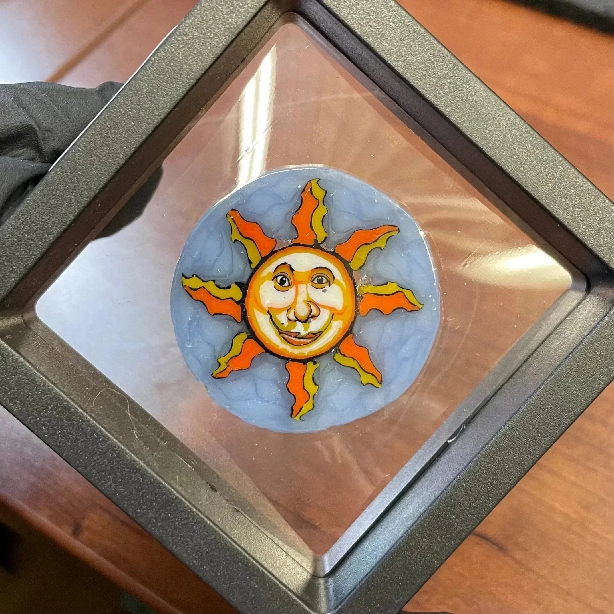 Handmade glass sun art with smiling face, displayed in protective frame on wood surface