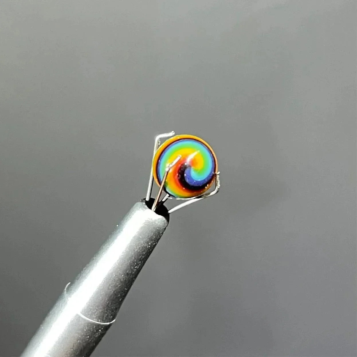 Colorful rainbow glass carb cap held by metal tweezers on a gray background