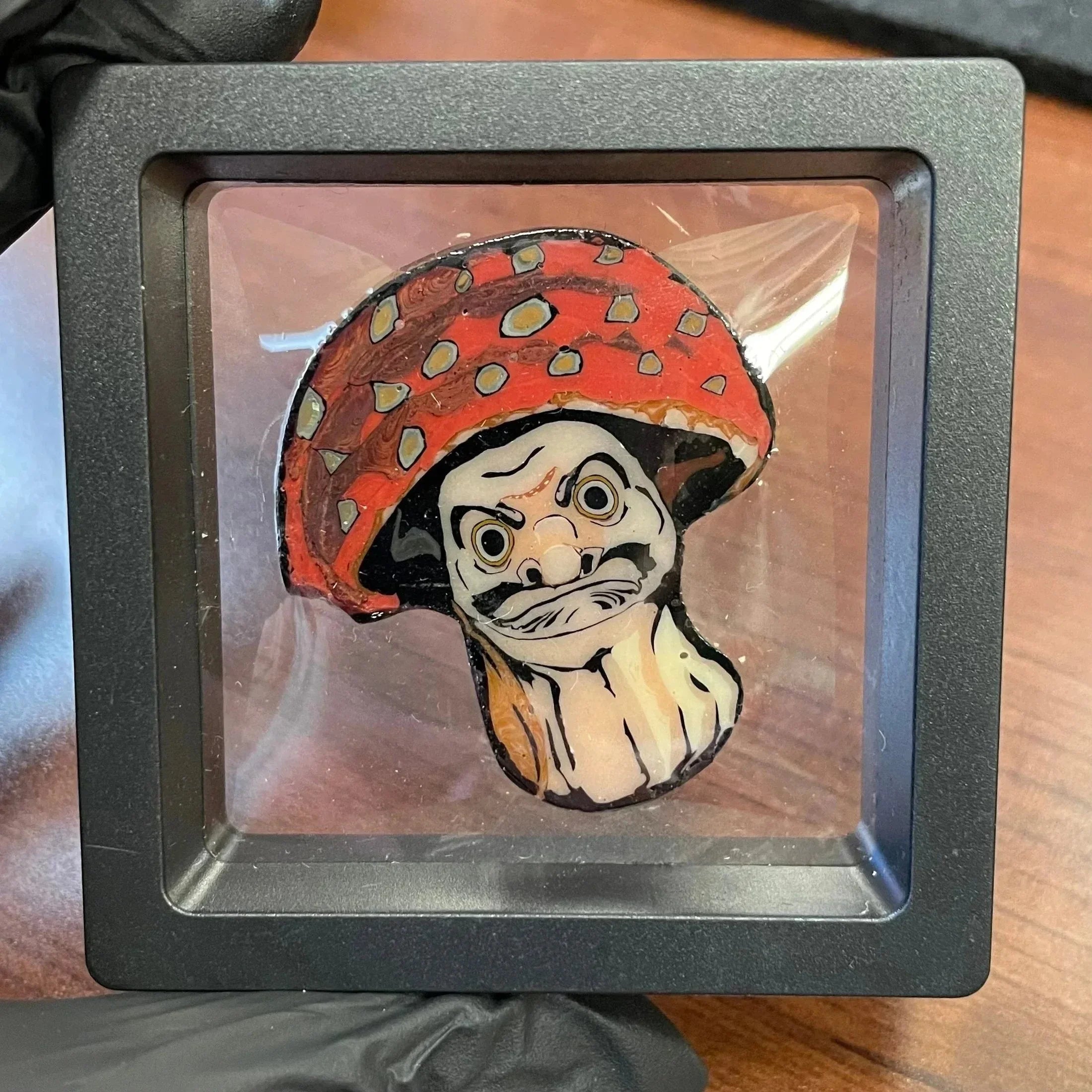 Handcrafted glass pin featuring a red mushroom with a cartoon face, displayed in a case.