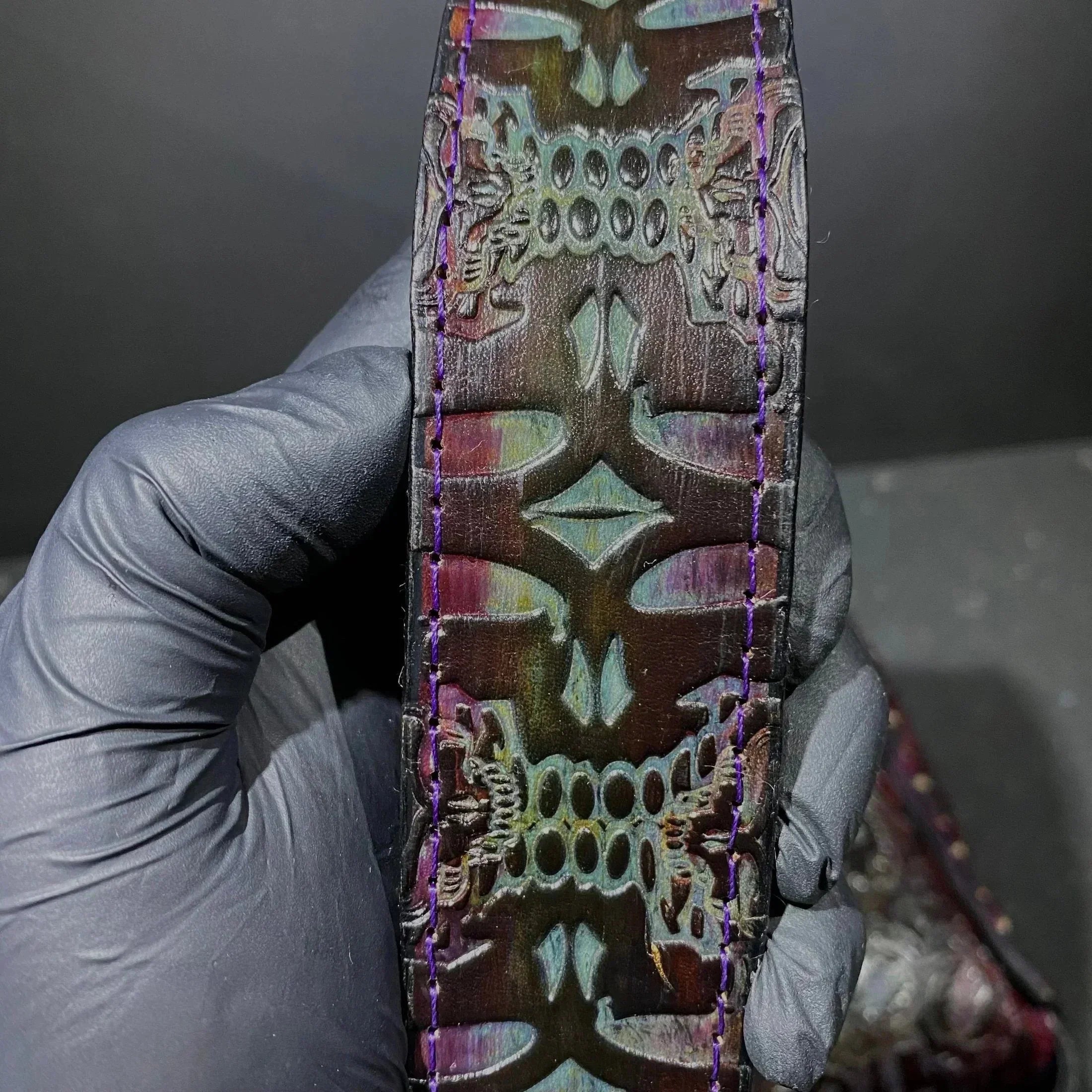 Hand holding carved iridescent leather strap with detailed pattern and purple stitching