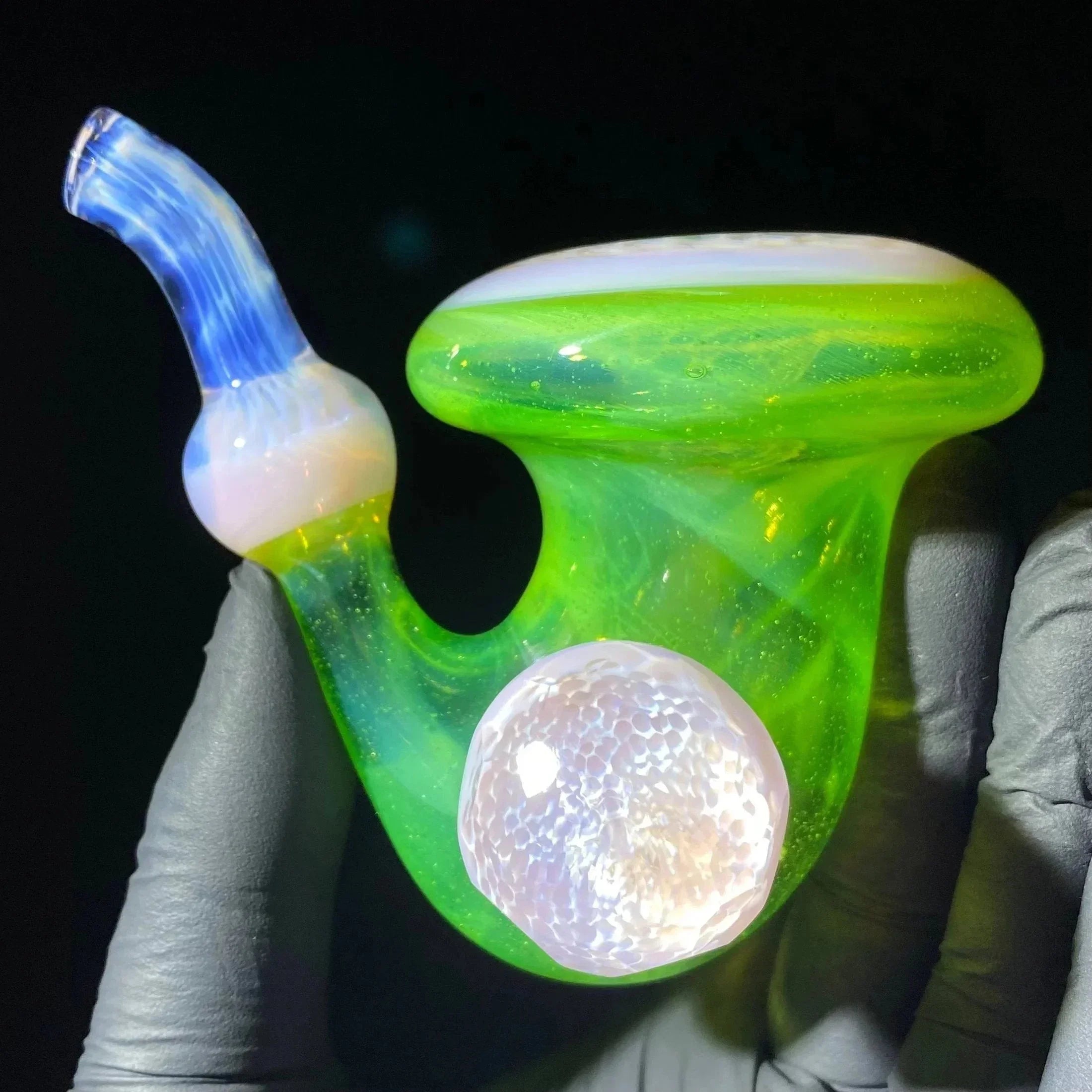 Hand holding a vibrant green and blue heady glass pipe with opal accent, against black background.