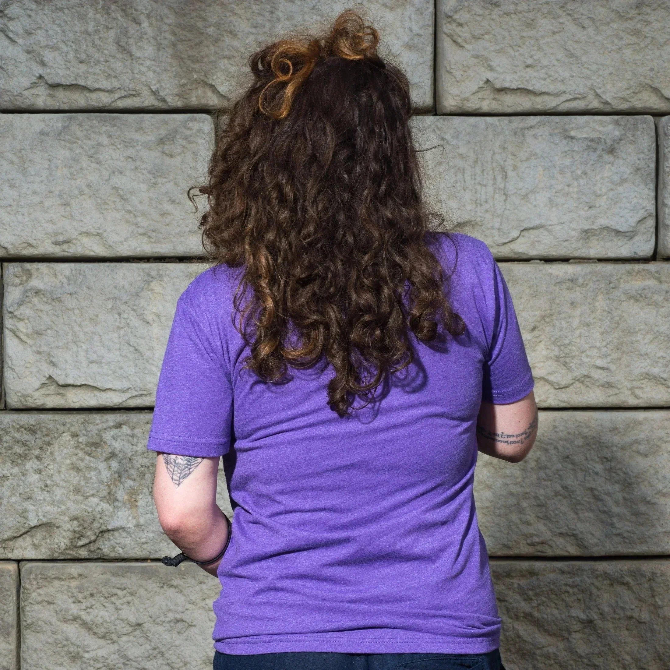 Woman with curly hair wearing purple t-shirt, standing against stone wall, showcasing casual style