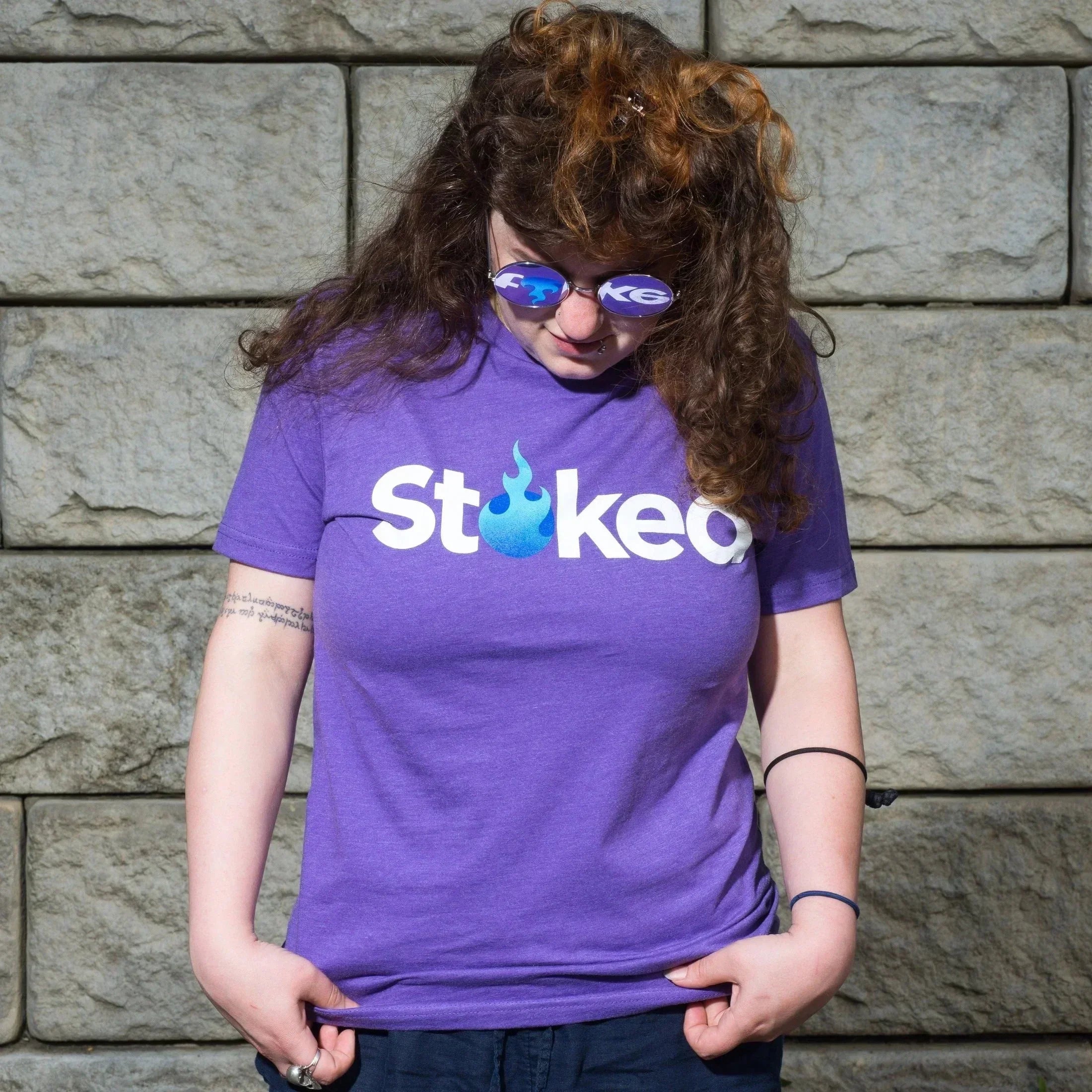 Purple Stoked logo t-shirt with blue flame design, worn by person with curly hair against stone wall