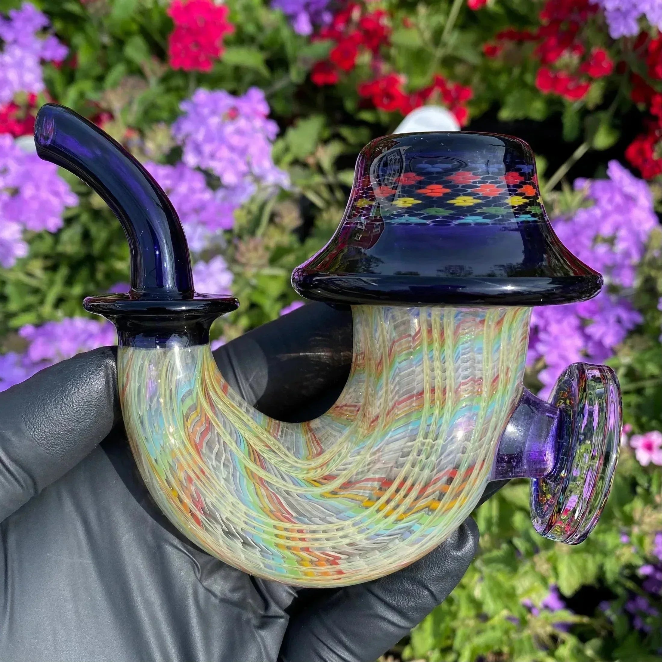Colorful heady glass pipe with intricate patterns held outdoors, purple and red flowers in background