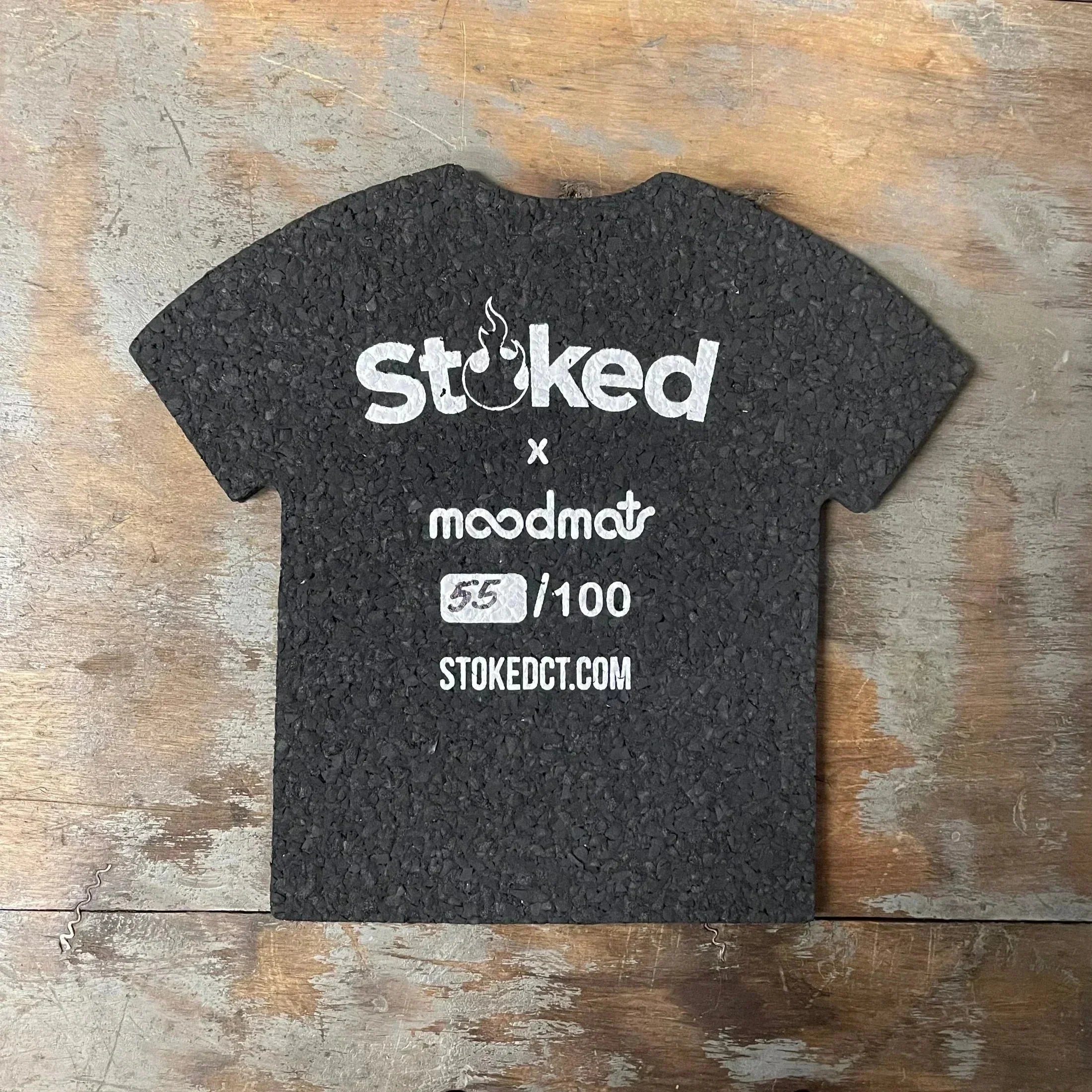 Black t-shirt shaped mat with white 'Stoked x moodmats' text, limited edition 55/100, on wood surface.
