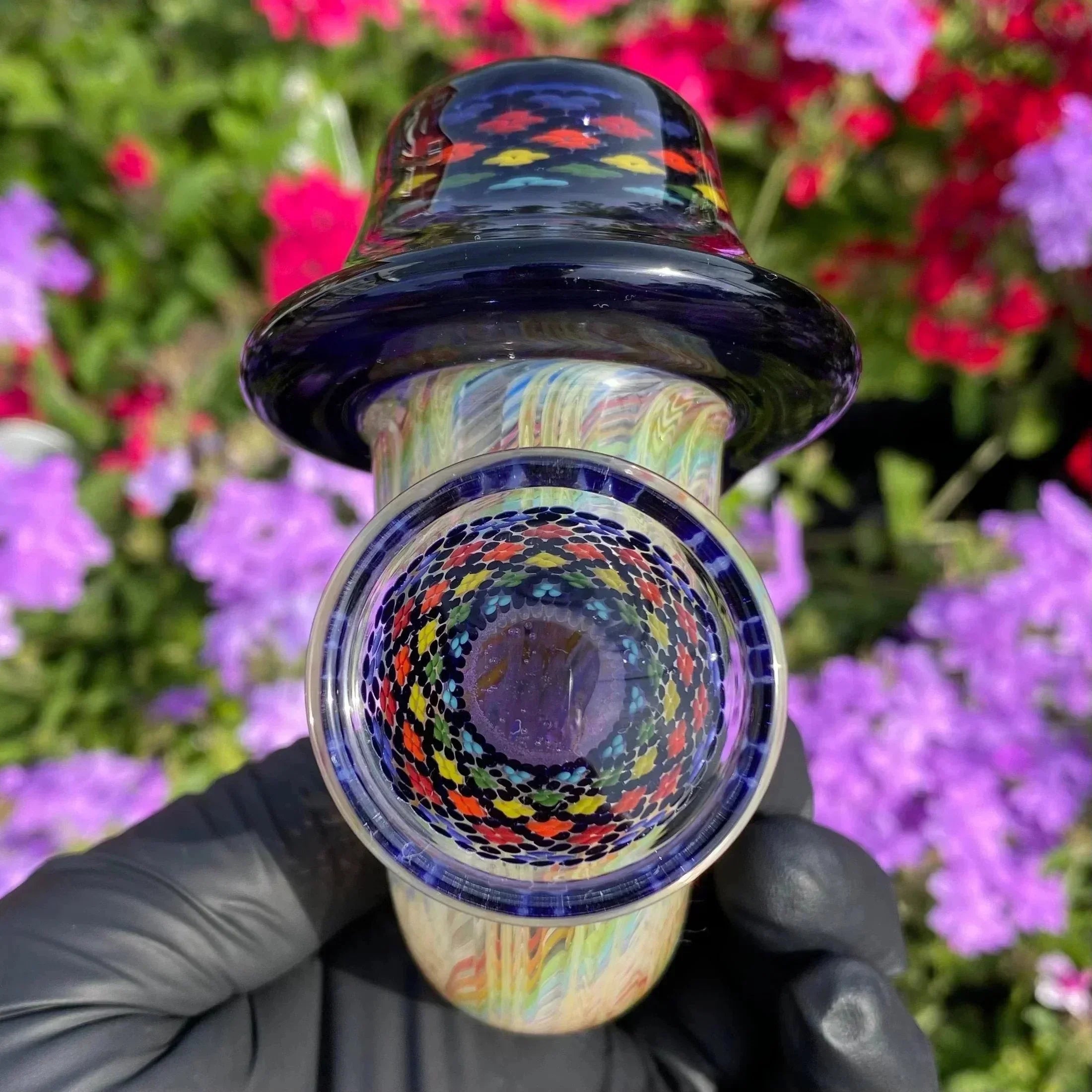 Colorful glass hand pipe with intricate pattern, held outdoors in front of purple flowers