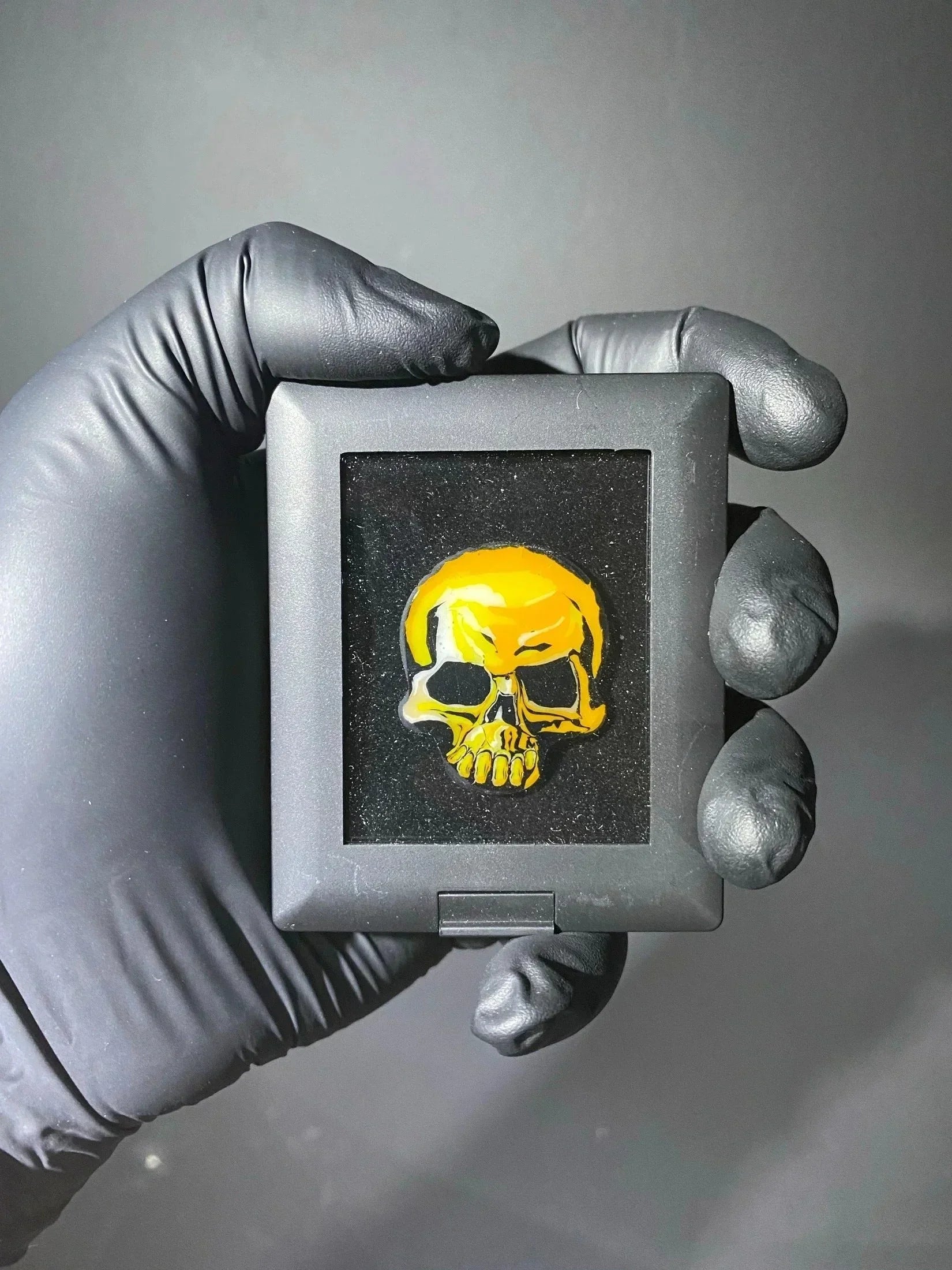 Hand holding a display case with a yellow glass skull pendant, black glove, dark background