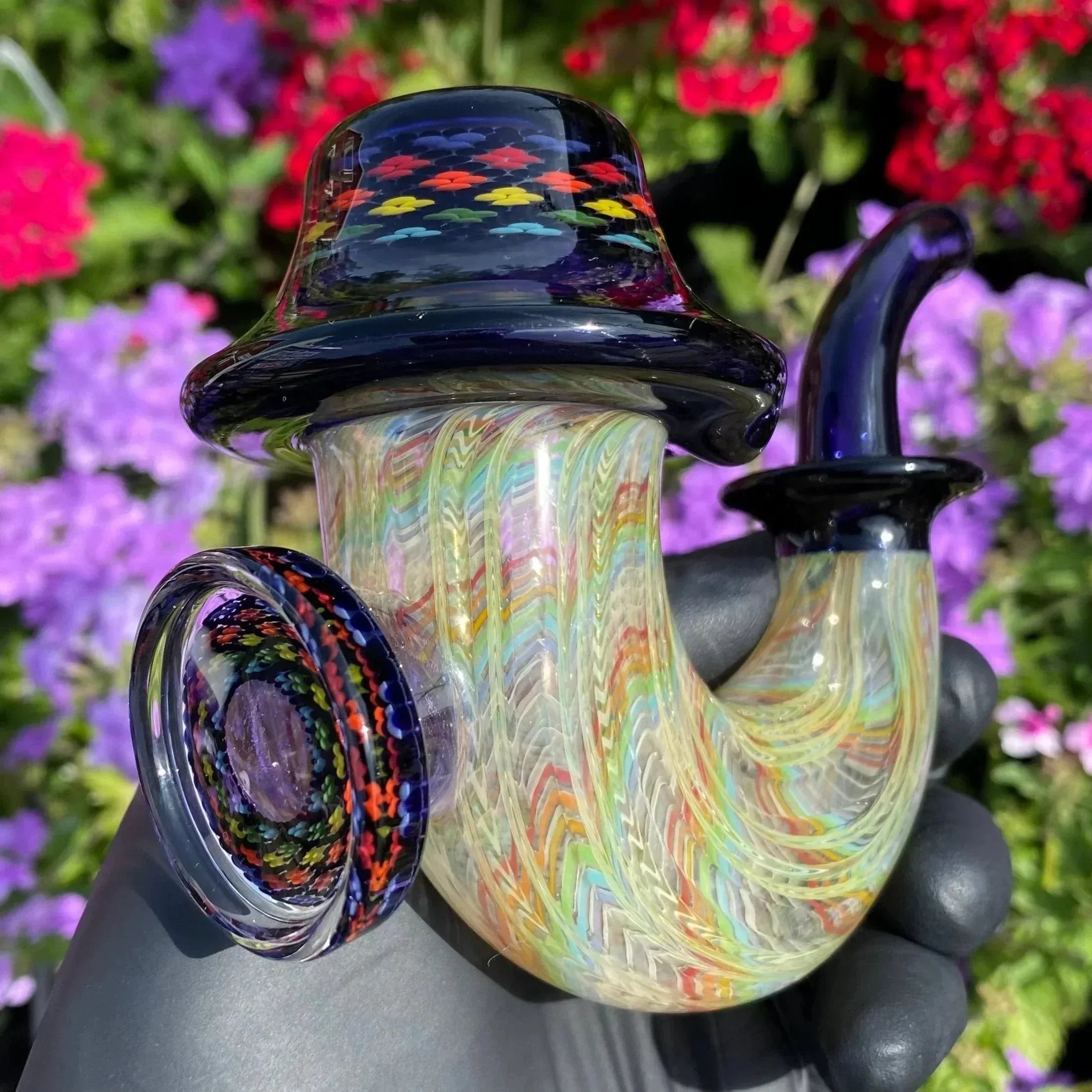 Colorful hand-blown glass pipe with intricate patterns, held outdoors by a gloved hand, flowers in background.