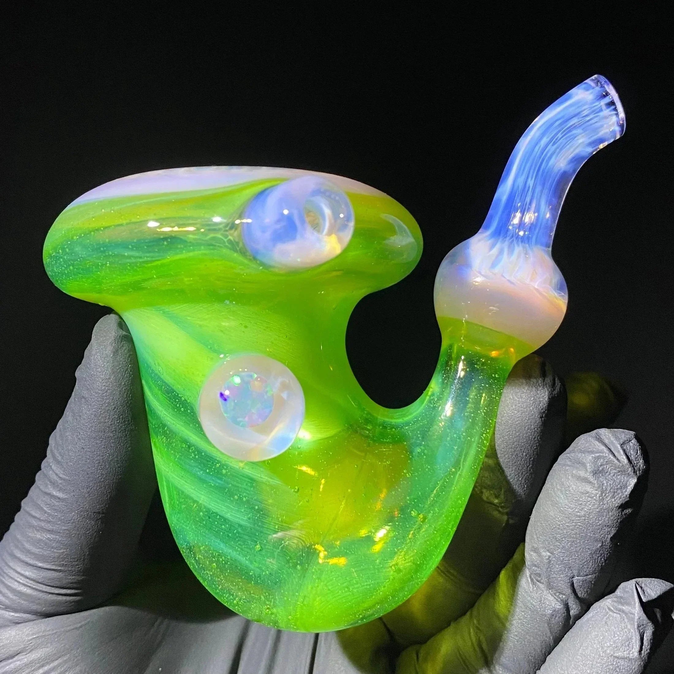 Hand holding a vibrant green and blue heady glass pipe with opal accents against a black background