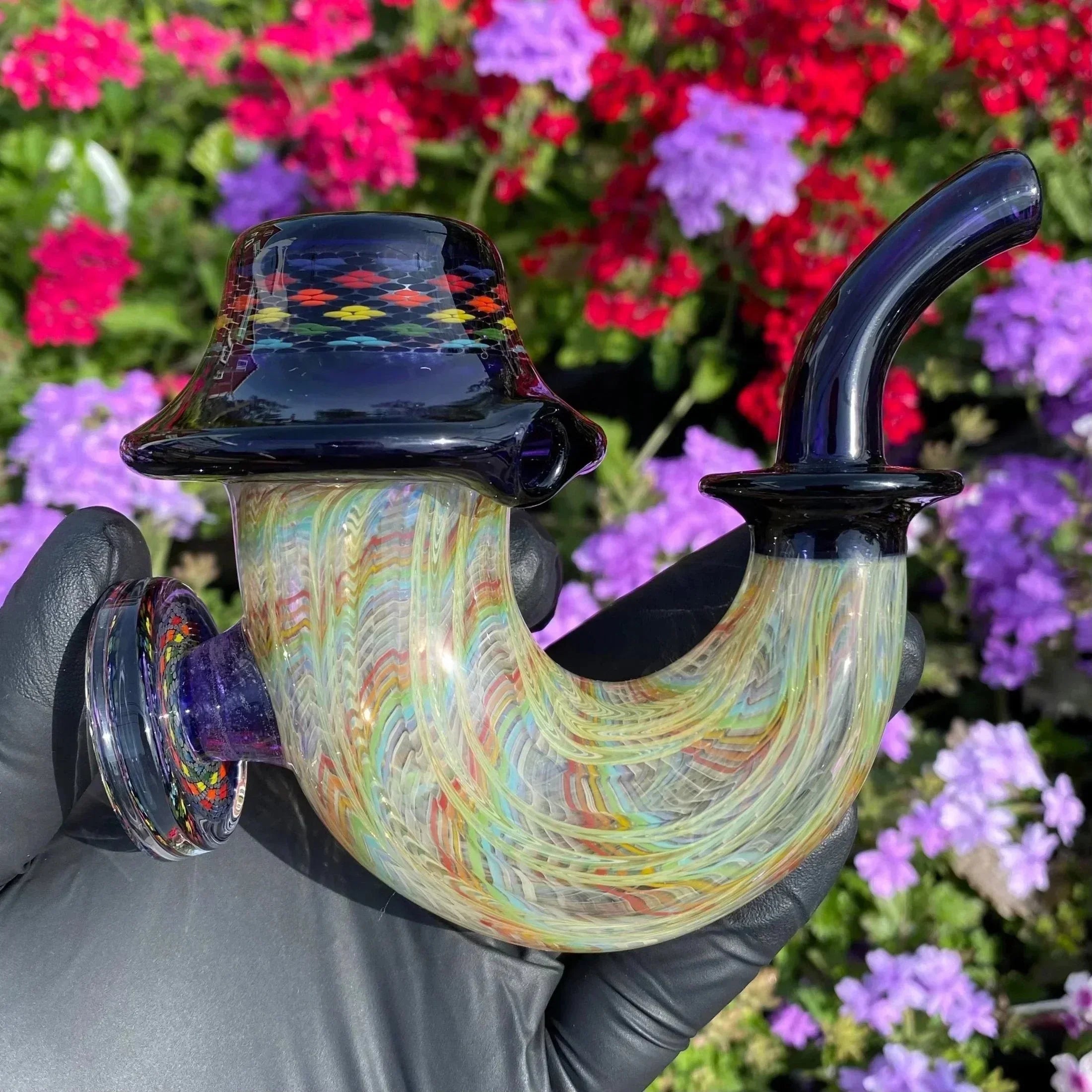 Colorful heady glass pipe with intricate patterns, held outdoors with flowers in background