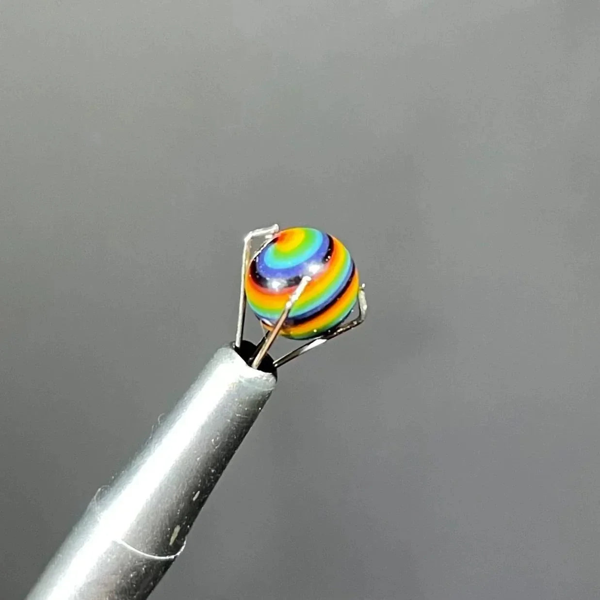 Close-up of a rainbow-striped glass marble carb cap held by metal tweezers