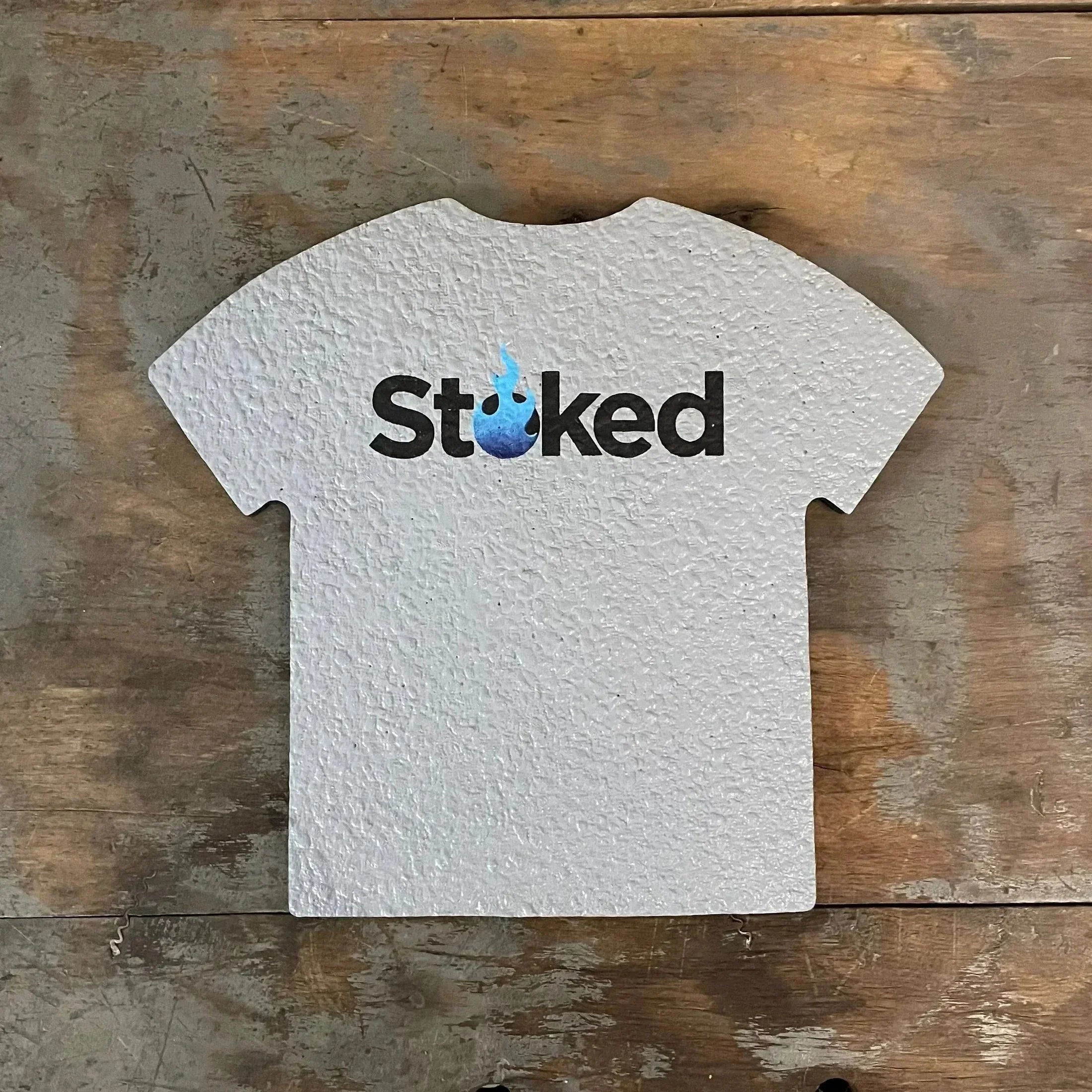 White t-shirt-shaped sign with 'Stoked' logo and blue flame, on rustic wood surface