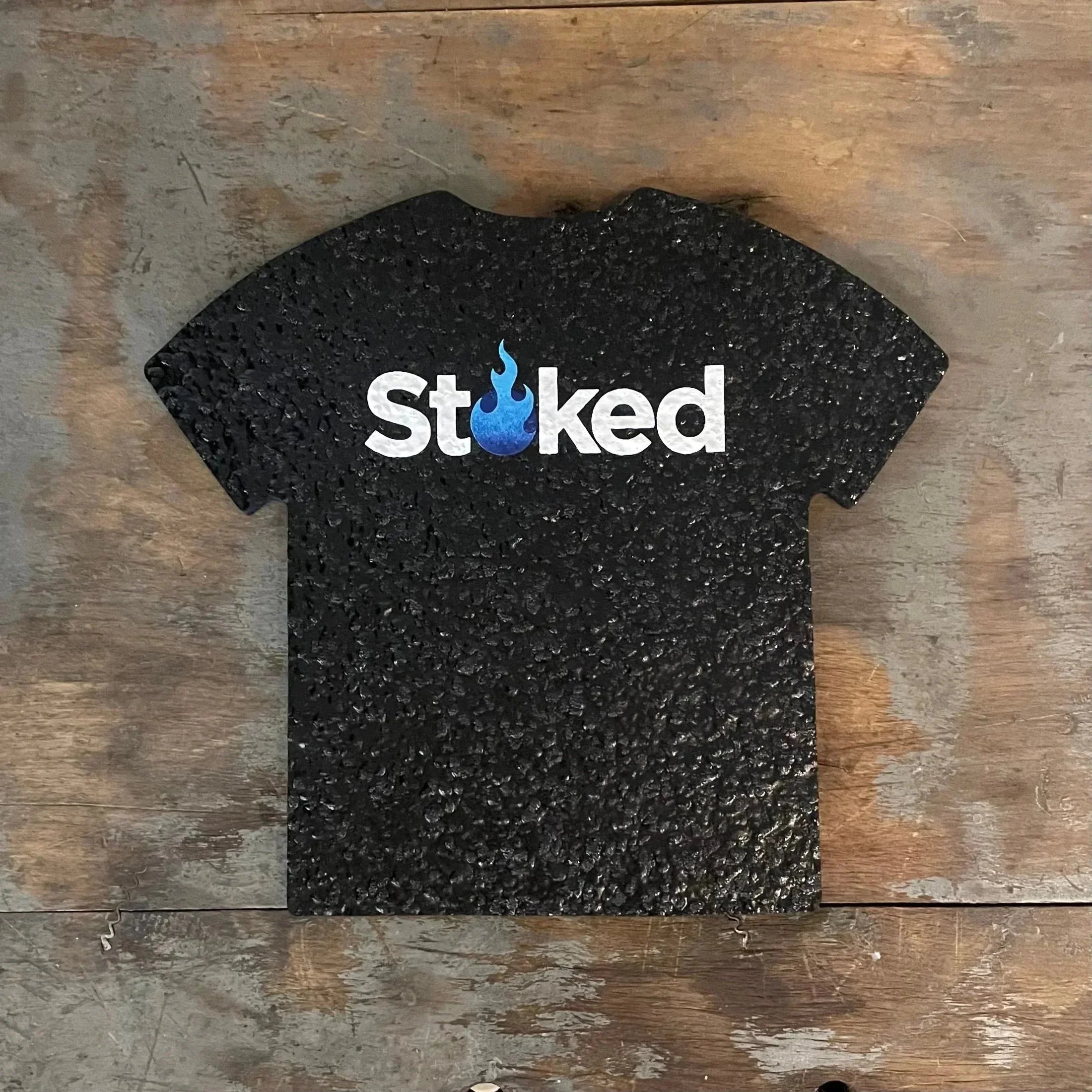 Textured black t-shirt display shaped like a shirt with 'Stoked' and blue flame graphic on wood surface.