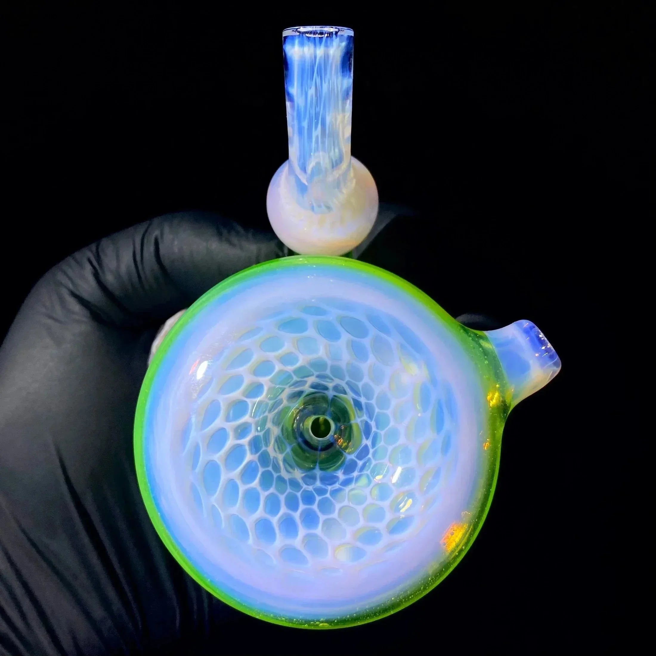 Hand holding a heady glass pipe with honeycomb pattern and blue, green accents on black background