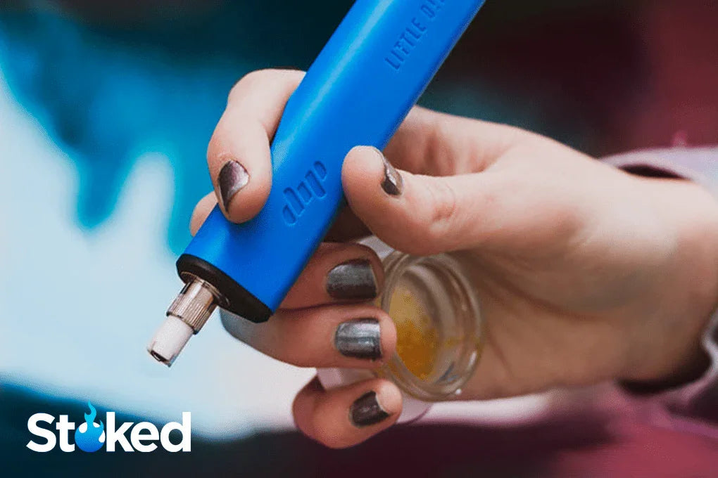 Hand holding a blue dab pen and small glass container with cannabis concentrate.
