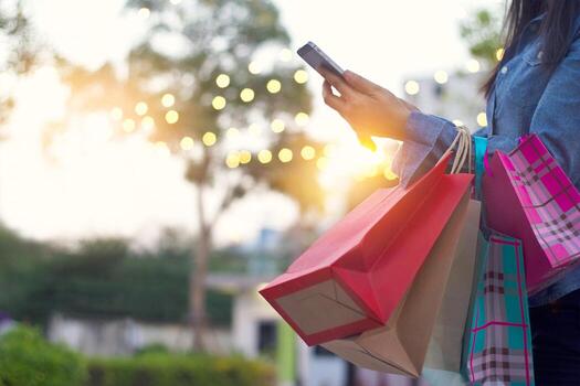 Woman holding shopping bags and using smartphone outdoors at sunset