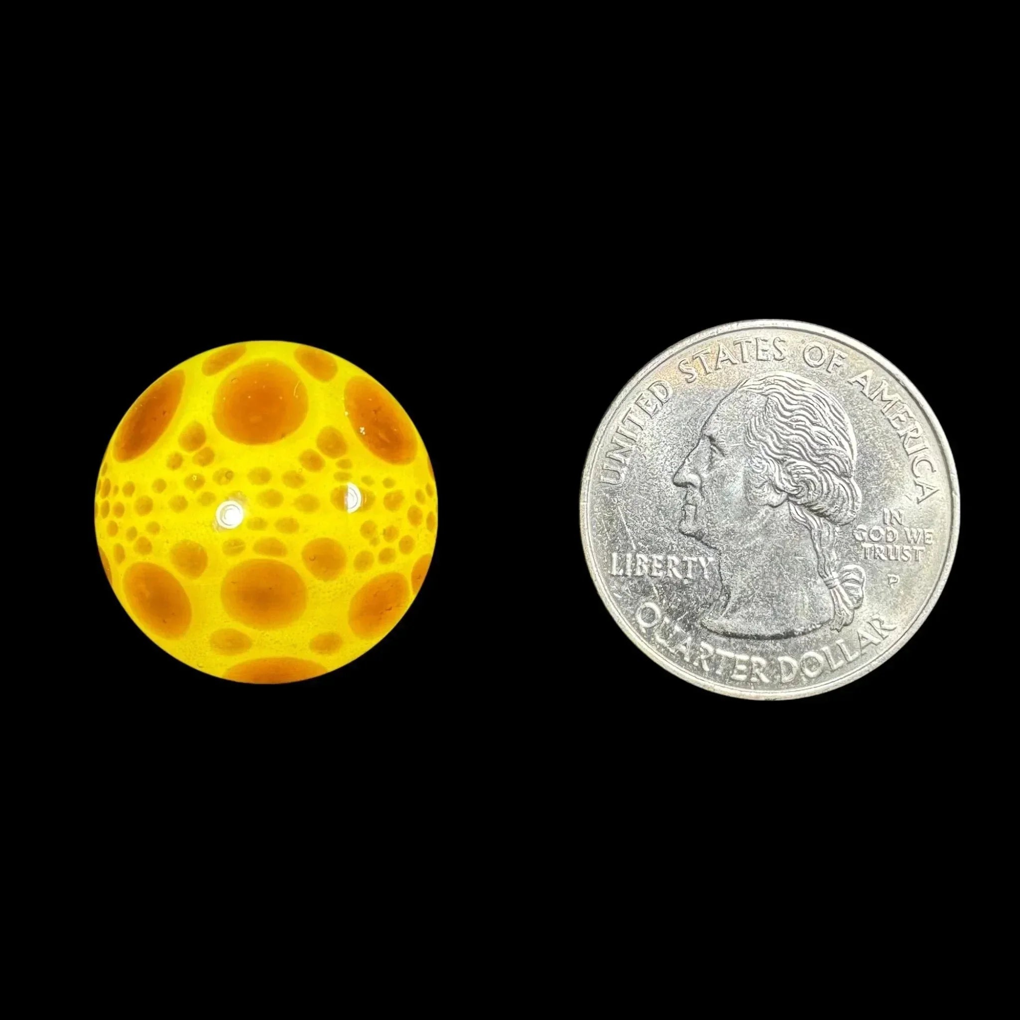 Yellow glass marble with orange crater pattern next to a US quarter on black background