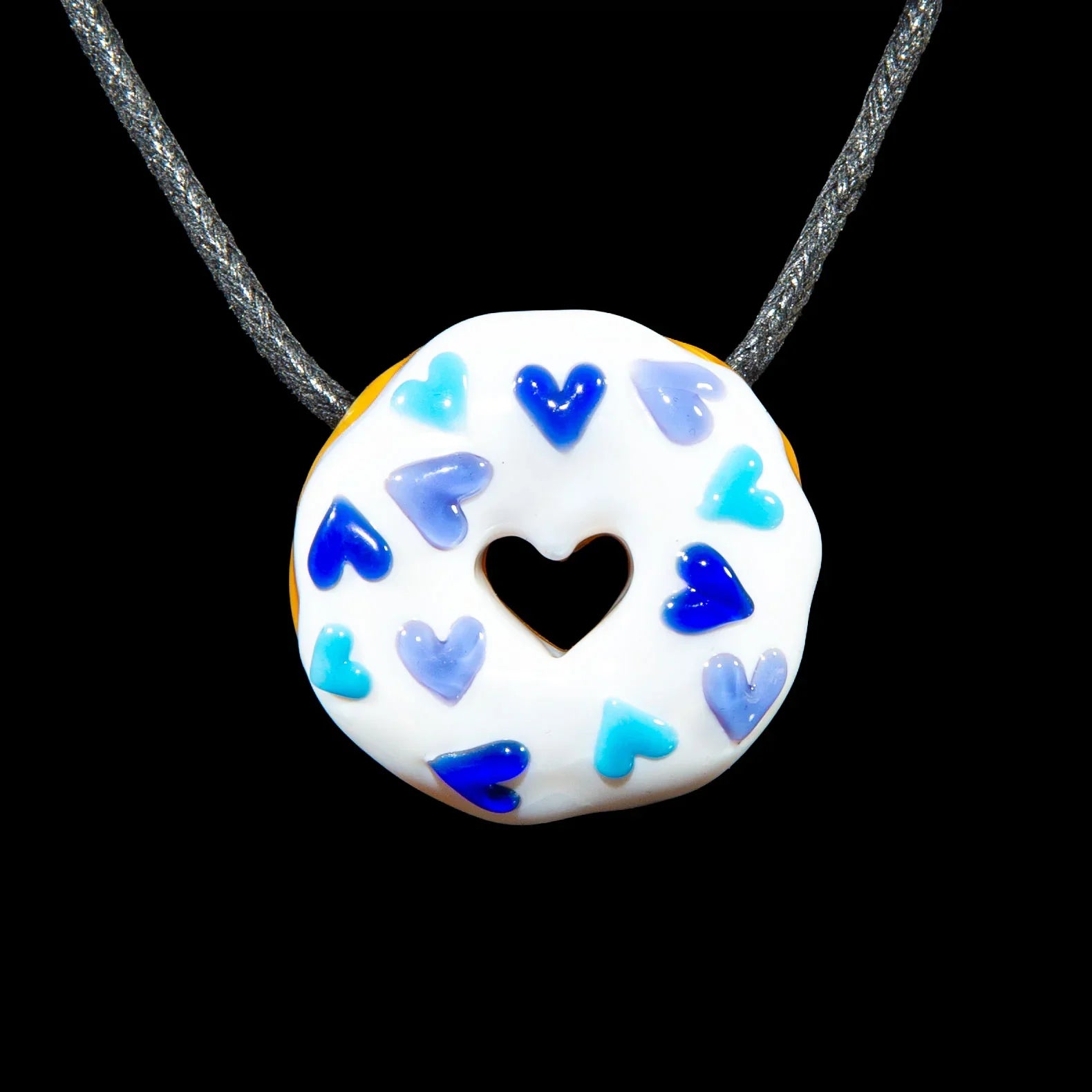 Glass donut pendant with white glaze, blue heart sprinkles, heart-shaped center, on cord
