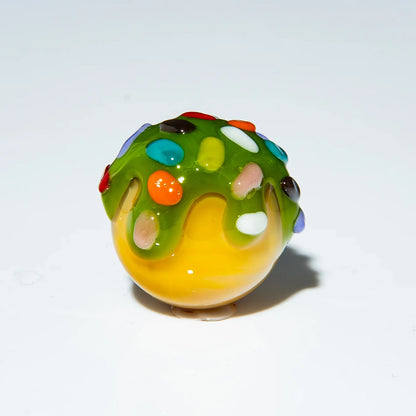 Handblown glass marble with yellow base, green drip design, and colorful sprinkle accents.