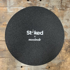 Stoked 17 inch circular mood mat on wood background with Stoked x Moodmats logo centered in white