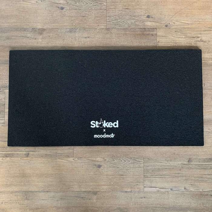 17x34 inch black moodmat on wood background, with Stoked x Moodmats logo centered bottom