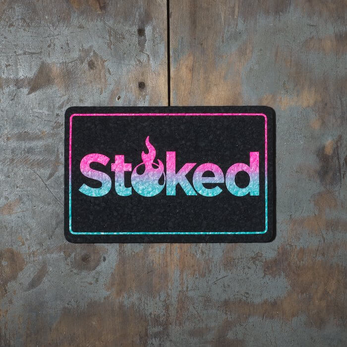 Stoked 8x5 inch black mood mat on a wood background. Stoked logo and outer outline are pink fading to teal.