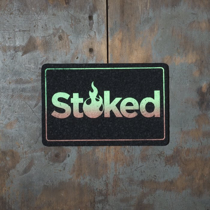 Stoked 8x5 inch black mood mat on a wood background. Stoked logo and outer outline are green fading to brown.
