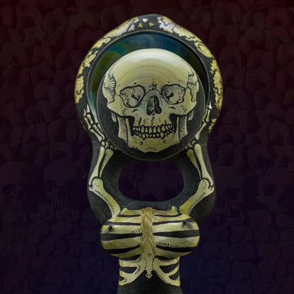 Hand-blown glass pipe with intricate skull and skeleton design on dark background